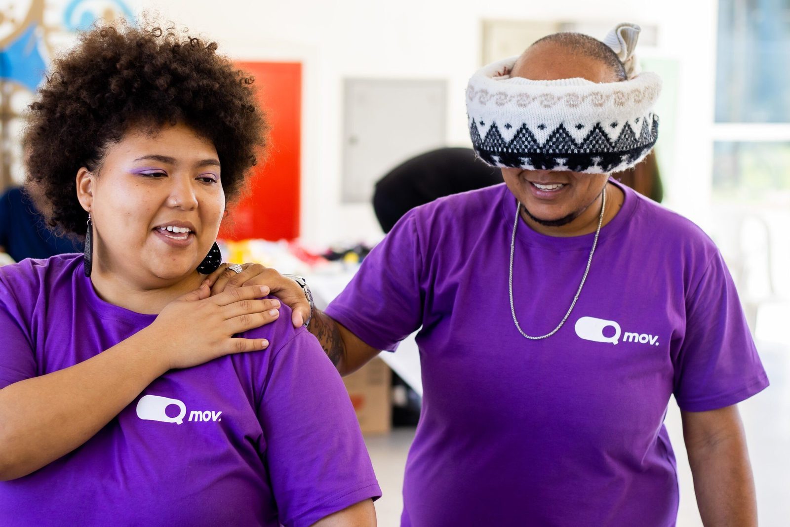 Dois participantes de uma atividade interativa vestem camisetas roxas com o logotipo ‘Q Mov.’. A pessoa à esquerda, uma mulher com cabelo crespo volumoso e maquiagem roxa, sorri enquanto guia a pessoa à direita, que usa um gorro puxado sobre os olhos como uma venda. Ela apoia a mão no ombro do colega, que sorri enquanto segura suavemente seu ombro. O ambiente parece ser um espaço interno iluminado, possivelmente um evento ou oficina.