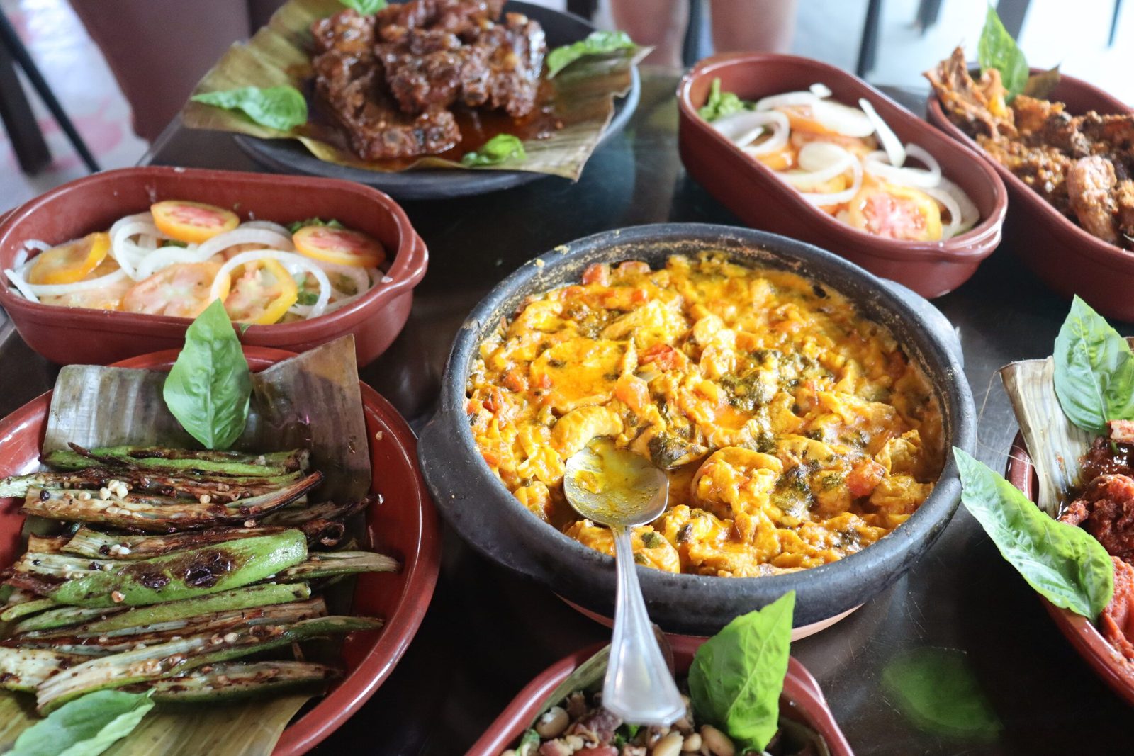 Mesa posta com destaque para panela de barro escuro com moqueca de caju dentro. A moqueca tem uma cor amarelada e a panela está com uma colher dentro.