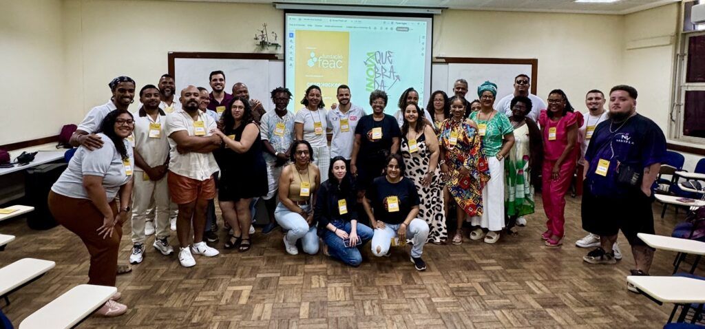 Registro coletivo de um encontro formativo realizado em uma sala universitária. A imagem reúne dezenas de participantes posicionados para uma foto em grupo, em um ambiente marcado pela diversidade racial, geracional e estética. Ao fundo, um projetor exibe a identidade visual do evento, reforçando o caráter colaborativo e comunitário da atividade. A composição transmite acolhimento, troca de saberes e construção coletiva.
